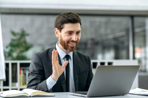  Man in suit sitting at a desk with an open laptop, waving at the people at the screen, during a video prospecting call